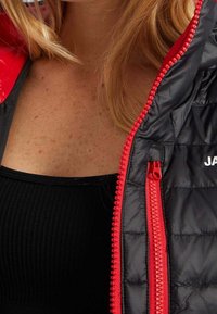 Veste isolante noire avec accents rouges et fermetures éclair, présentant un motif matelassé. Gros plan sur la zone de la fermeture éclair et le détail du col.