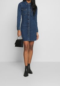 Denimskjortklänning i mörkblått, med långa ärmar, knappstängning fram, två bröstfickor och en figurnära silhuett. Bärs med svarta ankelskor.