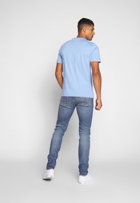 Vyras, dėvintis šviesiai mėlyną marškinėlį, liesas mėlynus džinsus ir baltus sportbačius, stovi ir žiūri į dešinę prieš paprastą baltą foną.