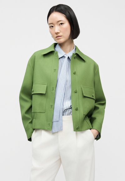 Femme aux cheveux courts et noirs portant une veste verte boutonnée sur une chemise rayée bleue et un pantalon blanc, debout les mains dans les poches.