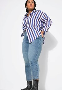 Camisa de botones a rayas azul y blanca con un corte holgado, combinada con unos vaqueros ajustados de un azul claro y botas negras de tobillo con acentos decorativos.