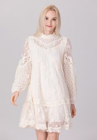 Robe en dentelle crème avec des manches bouffantes, des motifs floraux et un ourlet froncé. Présente un col haut et des détails transparents délicats.