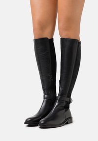 Bottes en cuir et daim noires montantes jusqu'au genou, avec une fermeture éclair sur le côté, un accent de boucle et un talon plat. Texture lisse avec un design épuré.