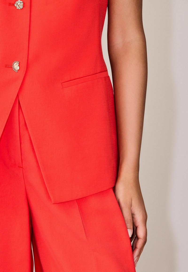 Lähivõte inimesest, kes kannab punast pükste komplekti kuldsete dekoratiivsete nuppudega, keskendudes torsole ja paremale käele.