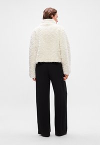 Λευκό, αφράτο κοντό μπουφάν με ψηλό γιακά, συνδυασμένο με φαρδιά μαύρα παντελόνια, με λεία υφή και χωρίς ορατά αξεσουάρ.