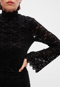 Μαύρη δαντελένια μπλούζα με ψηλό λαιμό και μακριά καμπάνα μανίκια. Διαθέτει λουλουδάτα μοτίβα, υφή υφάσματος και εφαρμοστή σιλουέτα.