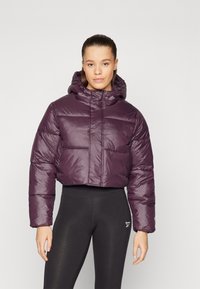 Giacca puffer cropped viola con cappuccio, caratterizzata da una texture lucida e trapuntata e polsini elastici. Abbinata a leggings neri.