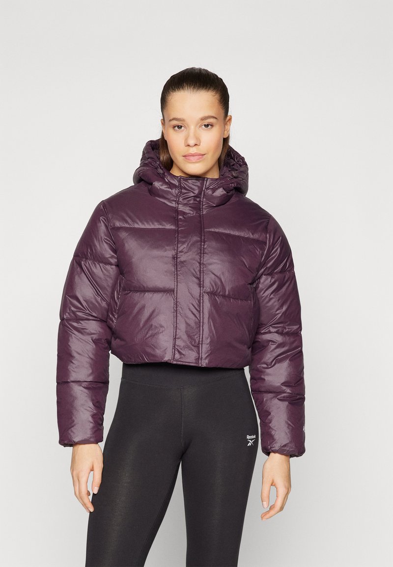 Giacca puffer cropped viola con cappuccio, caratterizzata da una texture lucida e trapuntata e polsini elastici. Abbinata a leggings neri.