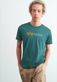 Camiseta de algodón en color verde azulado con cuello redondo, presenta un logotipo dorado y el texto "ALPHA INDUSTRIES" en la parte frontal, combinada con pantalones beiges.