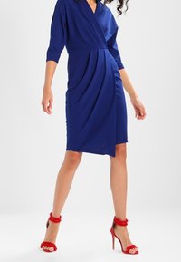 Vestido envolvente azul con mangas tres cuartos, diseño drapeado y corte a la altura de la rodilla. Combinado con tacones de aguja rojos con acentos de piel.
