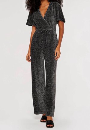 Kvinde iført en sort glitrende slå-om-jumpsuit med korte ærmer, vide ben og bælte i taljen, kombineret med sorte åbne sandaler.