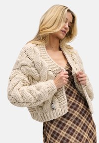 Beżowy, gruby, dziergany kardigan z wzorami warkoczy, szerokimi rękawami i krótkim fasonem. W połączeniu z brązową, kratką sukienką pod spodem.