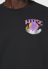 T-shirt en coton noir avec un design graphique comprenant le texte « NIKE » en rose et violet, un ballon de basketball coloré, et des accents de nuages à l'avant.