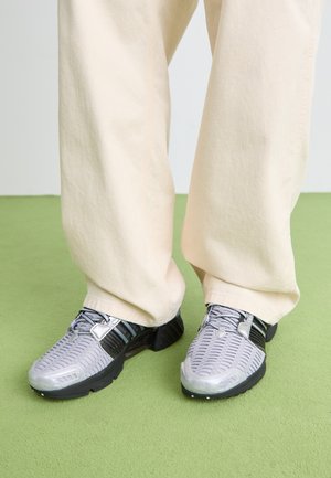 Person in beigen, weit geschnittenen Hosen und grauen Sport Sneakers mit schwarzen Sohlen steht auf einem grünen Teppich.