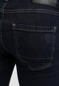 Acercamiento del bolsillo trasero de unos pantalones vaqueros de mezclilla azul oscuro con costuras amarillas y un pequeño logotipo bordado redondo en el bolsillo.