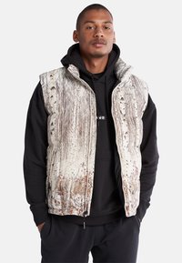 Isolierte Weste mit einem von der Natur inspirierten Design, das eine cremefarbene Basis und braune Baum-Muster aufweist, mit Reißverschluss vorne, hohem Kragen und Seitentaschen.