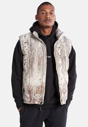 Gilet isolant avec un design inspiré de la nature, présentant une base crème et des motifs d'arbres marron, fermeture éclair à l'avant, col haut et poches latérales.