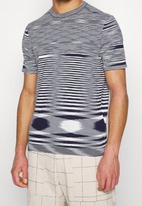 Kortärmad stickad topp med ett horisontellt randmönster i grått, marinblått och vitt, med rund hals och texturerad yta.