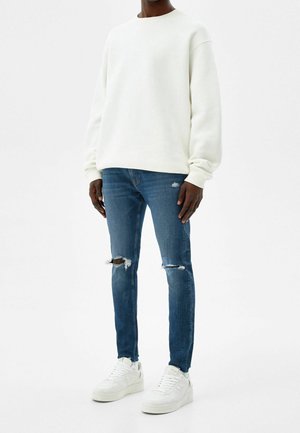 Felpa bianca dalla vestibilità comoda, abbinata a jeans blu a taglio slim con dettagli consumati. Completano il look le sneakers bianche.