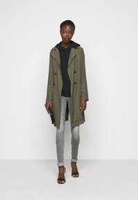 Casaco trench de duplo peito verde oliva, com um hoodie cinza de malha por baixo, combinado com jeans cinzentos e sandálias de salto pretas; segurando uma clutch preta.