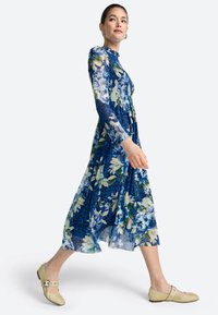 Obleka z dolgimi rokavi v modrem cvetličnem vzorcu, z oblikovanim zgornjim delom in tekočo krilno linijo. Model nosi obutev v svetlo rumeni barvi.