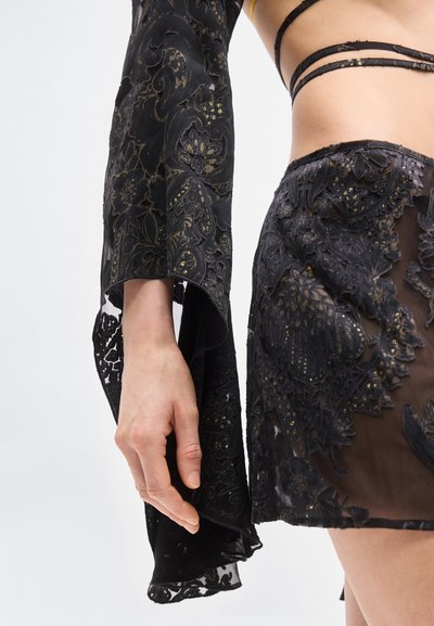Γυναίκα που φοράει διαφανή μαύρη δαντελένια φούστα και μακρυμάνικο τοπ με κεντήματα λουλουδιών και χρυσές λεπτομέρειες, με εμφανές το μέσο σώμα και το χέρι.
