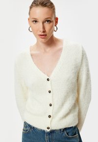 Cardigan bianco fluffy con scollo a V, maniche lunghe e bottoni marroni lungo il davanti. Il tessuto sembra morbido e texturizzato.