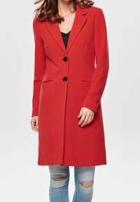 Abrigo rojo con solapas muescas, dos botones negros y bolsillos frontales. Textura suave, diseño ajustado, usado sobre una camiseta negra y jeans rasgados.