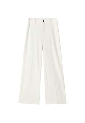 Kobieta stojąca z założonymi ramionami, ubrana w jasnoróżową czapkę, czarną kurtkę dżinsową, białe szerokie spodnie oraz czarne buty na beżowym tle.