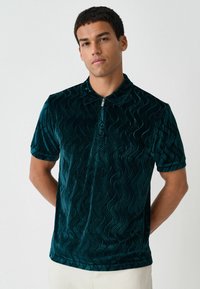 Tealowa welurowa koszulka polo z teksturowanym falistym wzorem, krótkimi rękawami i kołnierzykiem na pół zamek. Charakteryzuje się miękkim, błyszczącym wykończeniem.