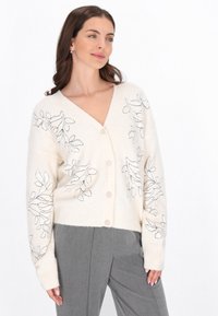 Cremfarbener Cardigan mit tiefem V-Ausschnitt, der schwarze gestickte Blättermuster auf der Vorderseite und gerippte Bündchen aufweist. Mittelschwerer Strickstoff.