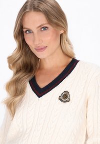 Cremfarbener Cable-Knit-Pullover mit einem dunkelblauen V-Ausschnitt und roter Streifen. Besitzt ein gesticktes Patch mit einer Krone. Weicher Stoff und klassischer Schnitt.