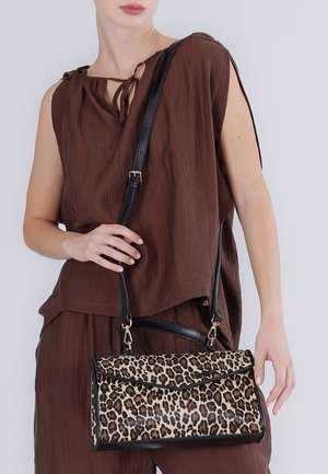 Mujer con top sin mangas marrón y pantalones, sosteniendo un bolso con estampado de leopardo y una correa negra sobre el hombro.