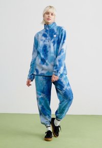 Blå tie-dye fleece-sett med høy krage, glidelås foran og matchende løse bukser. Myk tekstur med elastiske mansjetter og liv. 