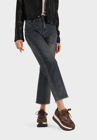 Donna che indossa jeans blu a vita alta con orli sfilacciati, sneakers platform marroni e una giacca di pelle nera, in piedi con una gamba incrociata.