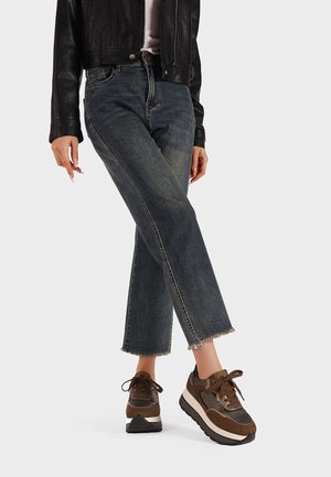 Donna che indossa jeans blu a vita alta con orli sfilacciati, sneakers platform marroni e una giacca di pelle nera, in piedi con una gamba incrociata.