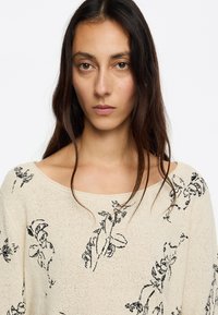 Pull en maille beige avec broderie florale noire, encolure large et manches longues. Tissu texturé avec une coupe ample et une silhouette décontractée.