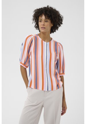 Kobieta o kręconych włosach, ubrana w zapinaną na guziki bluzkę w pionowe paski w kolorach pomarańczowym, różowym, fioletowym, białym i niebieskim, w połączeniu z beżowymi spodniami.