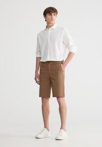 Camicia bianca leggera a maniche lunghe, pantaloni corti marroni con texture e sneaker bianche. I pantaloni corti presentano una vita e tasche laterali.