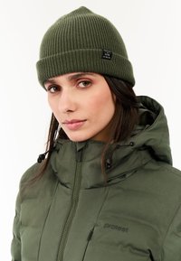 Groene geribbelde gebreide beanie met een opgevouwen rand en label. Draag het met een donkergroene gewatteerde jas met een hoge kraag en ritssluiting.