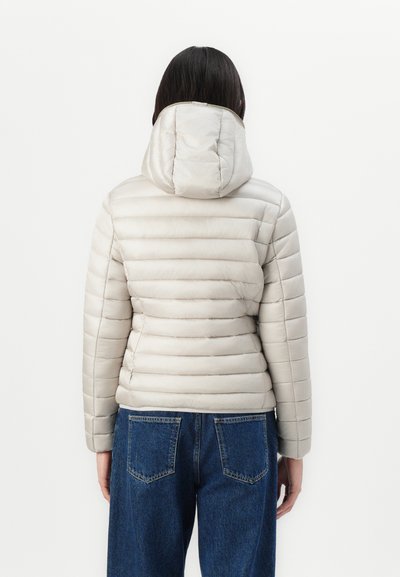 Veste matelassée beige clair avec capuche, présentant un design matelassé et une texture lisse. Portez-la avec un jean en denim bleu, montrant la vue arrière.