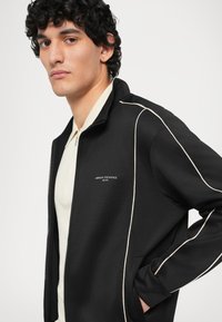 Chaqueta negra con cremallera y piping de color claro, que presenta dos bolsillos laterales y un pequeño logotipo en el pecho. Llevada sobre una camiseta polo color crema.