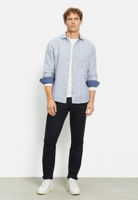 Camicia a maniche lunghe a righe blu e bianche con bottone frontale e polsini arrotolati, abbinata a pantaloni neri e sneakers bianche.