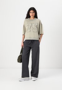 Överdimensionerad ljusgrön sweatshirt med broderad "75", kombinerad med mörkgrå vida byxor och vita sneakers. Inkluderar en liten grön väska.