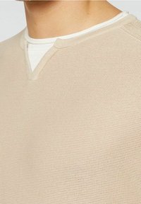 Pull en tricot beige avec un design en V, présentant une surface texturée et une chemise blanche superposée en dessous. Style simple et décontracté.