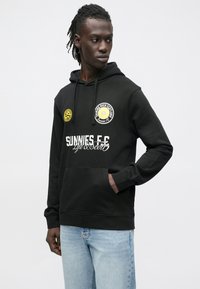 Sweatshirt preta com capuz, com vários patches impressos, incluindo um emblema amarelo e texto branco, com bolso canguru e punhos canelados.