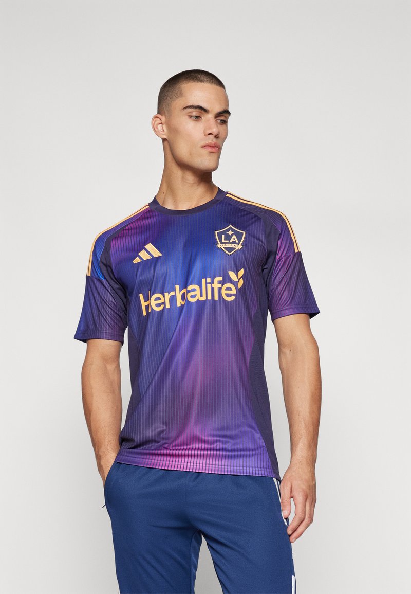 Maglia da calcio viola con strisce verticali, dettagli arancioni e logo "Herbalife". Maniche corte e scollatura rotonda.