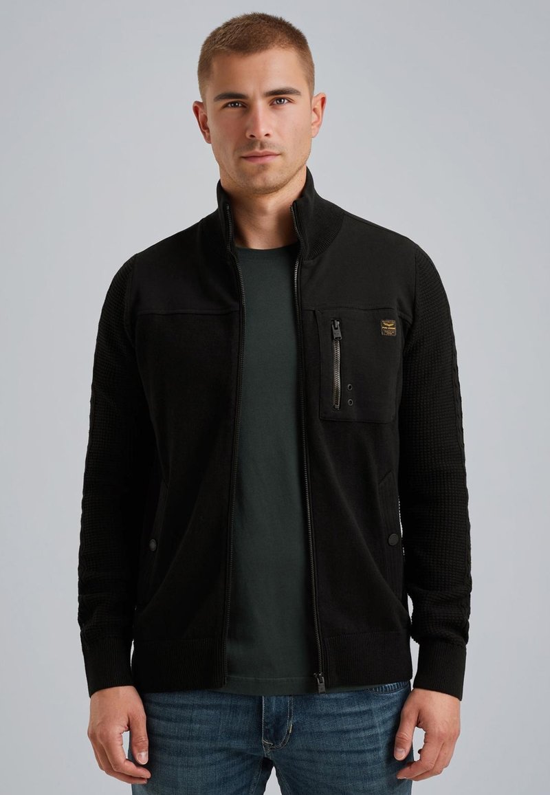 Musta vetoketjullinen takki tekstuurilla varustetuilla hihoilla, rintatasku vetoketjulla, pyöreä kaula-aukko ja istuva malli, käytetty tummanvihreän paidan päällä.