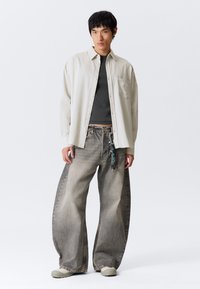 Young man wearing oversized light beige shirt, black cropped top, wide faded gray jeans with keychain, and beige shoes standing on white background.