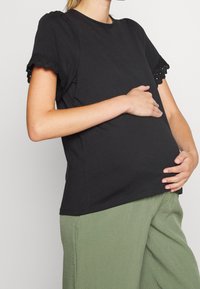 Svart bomullströja med vågformade spetsdetaljer på de korta ärmarna, figurnära silhuett, ihopparad med lösa gröna byxor.